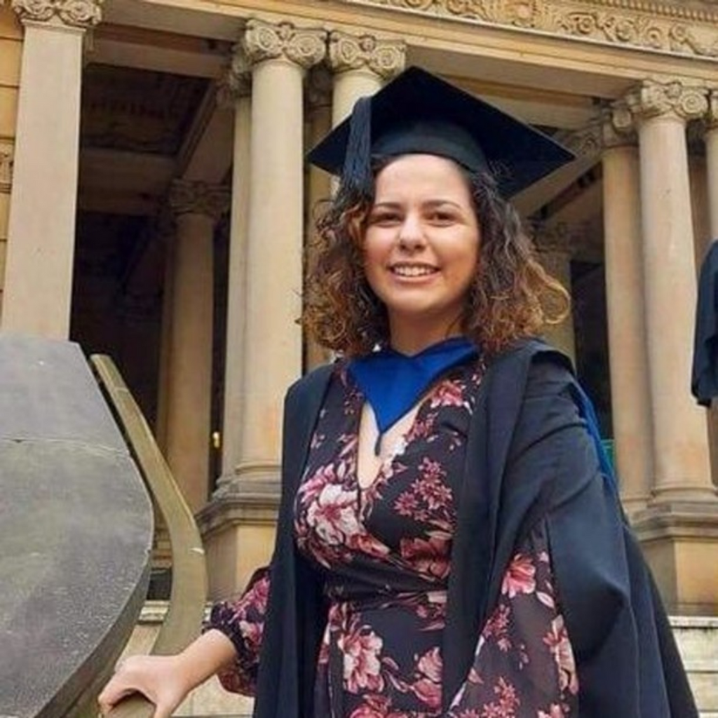 A photo of Alyssa in her graduation gown. She is wearing a black dress with pink florals. She is holding the hand railing of a staircase and smiling brightly at the camera. Her short brown to blind ombre hair is flowing freely. 