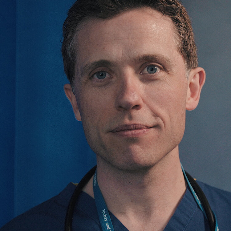 A close-up image of Dr Alan Desmond. He is wearing his scrubs and has a lanyard around his neck. He is lightly smiling and looking directly at the camera. 