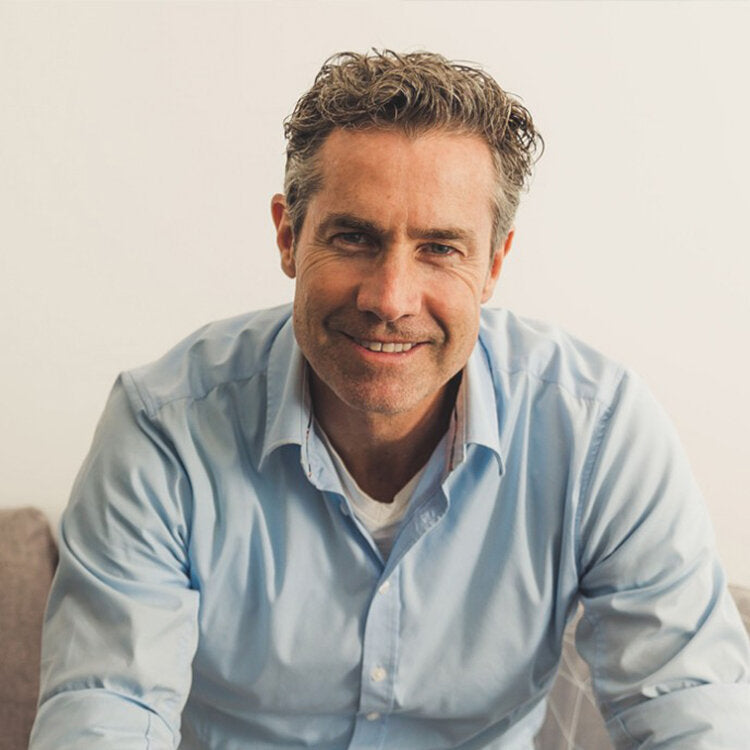 An image of Tom Cronin smiling into a camera. The image is close up but you can see he is sitting on a couch with his arms resting on his knees. He is slightly hunched over to position his face closer to the camera. He is wearing a pale blue button up