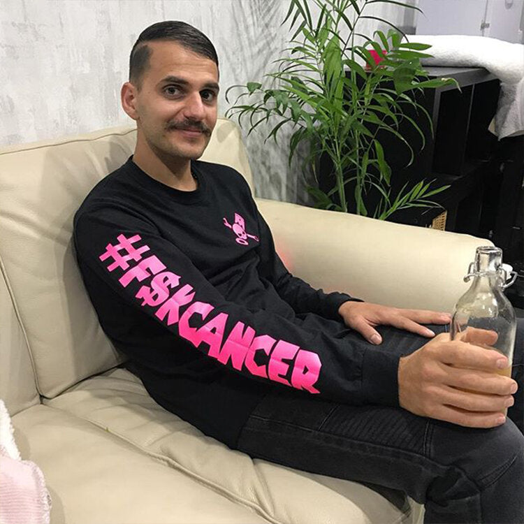 An image of Dom Tripodi sitting on a white leather couch. He is wearing an anti-cancer jumper and holding a glass jar filled with water. He is smiling at the camera. The background features a small indoor plant and a countertop.