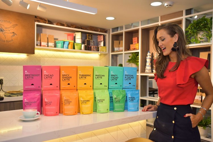 Vicki Nguyen, the founder of Jomeis Fine Foods standing in front of the nutritional latte collection. From left to right, the flavours are; beetroot, cacao, spicy ginger, turmeric, matcha, minty cacao and matcha and cacao. Vicki is smiling at the lattes 