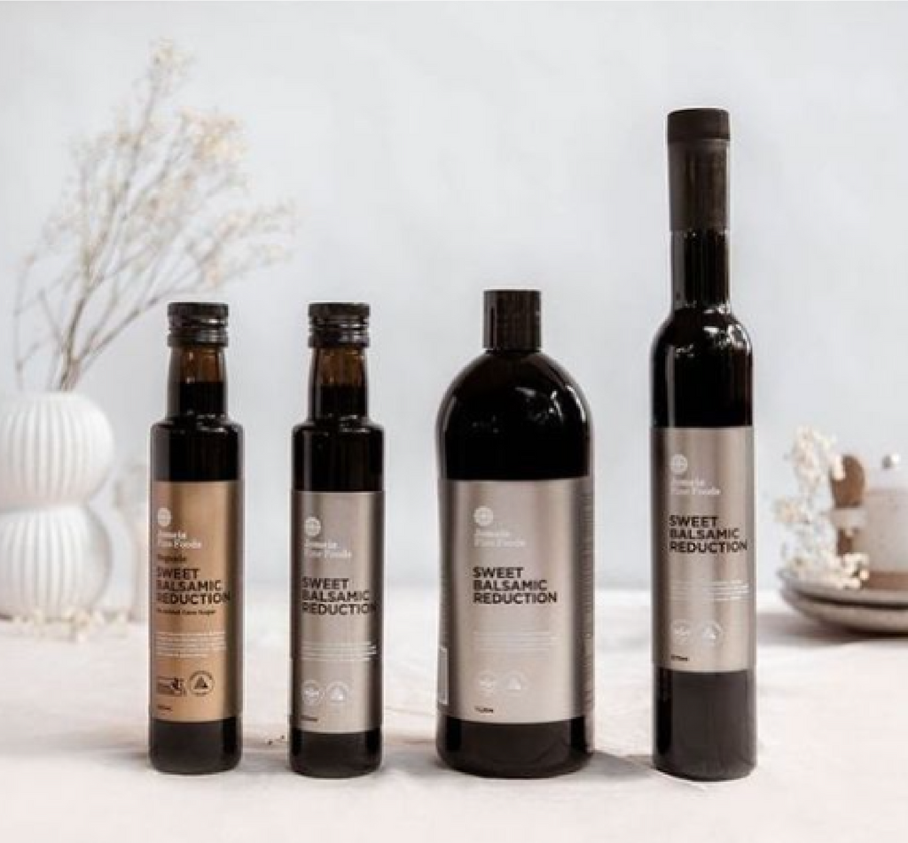 Four sweet balsamic reductions sitting in front of a delicate background. From left to right they are, organic 250 ml, regular 250ml, regular 1l and regular 350 ml. They all have silver logos aside from the organic which is gold.