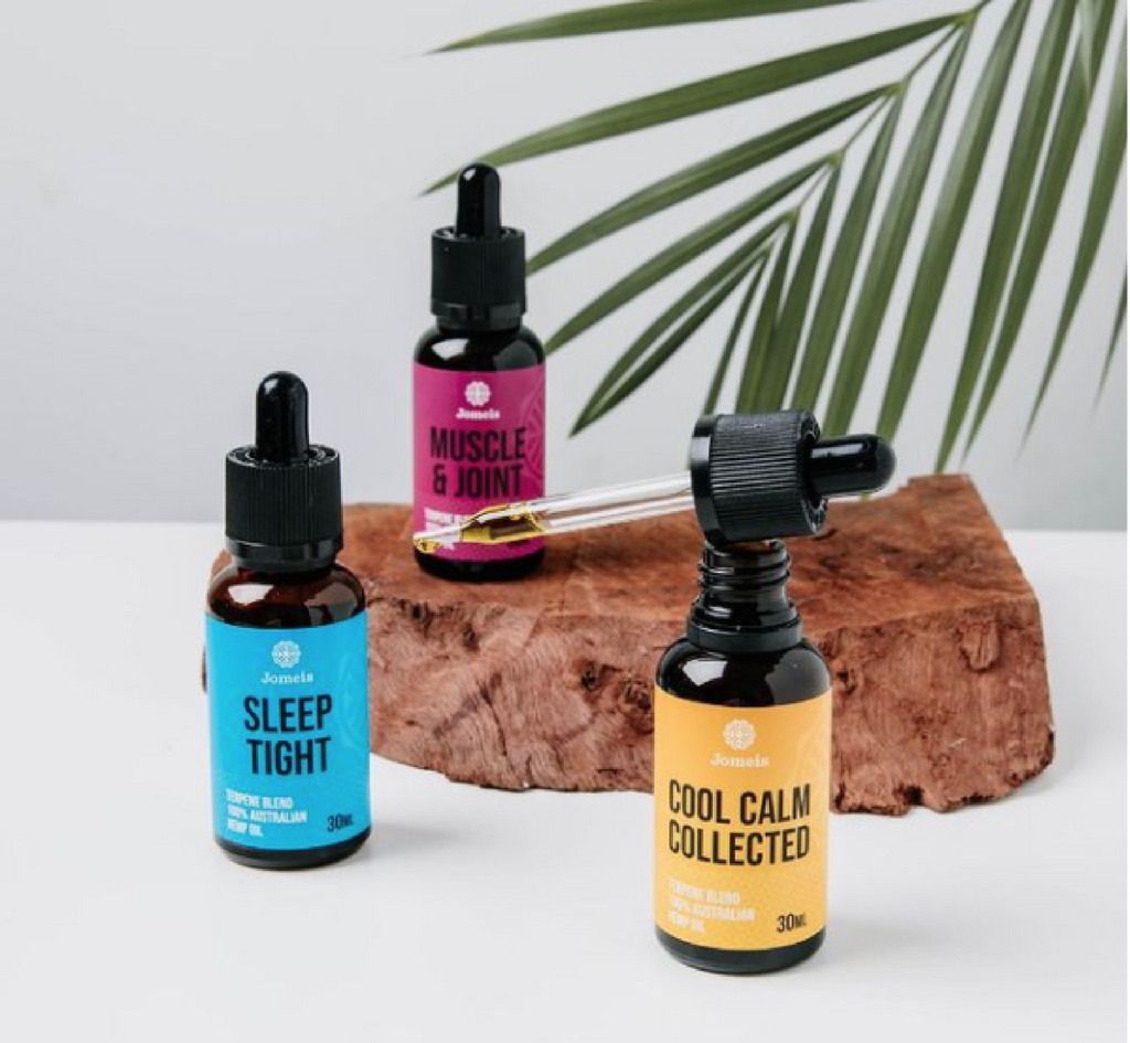 three terpene blends. Two of them are on a table while the third (a pink muscle and joint terpene) is sitting on a rock. behind the rock is a palm leaf. The terpene on the left is a blue sleep tight terpene the right is a yellow cool calm collected