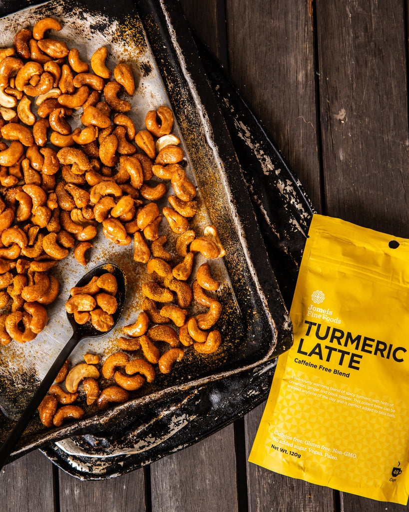 A turmeric latte packet laying on a wooden table. Beside it are roasted cashews on a baking tray which are coated in the turmeric powder 