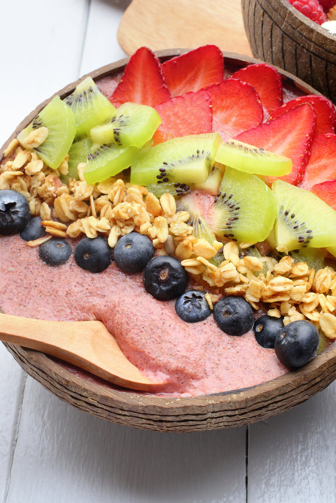 A superfood breakfast bowls topped with blueberries, oats, kiwi and strawberries. There is a wooden spoon in the breakfast bowl. 