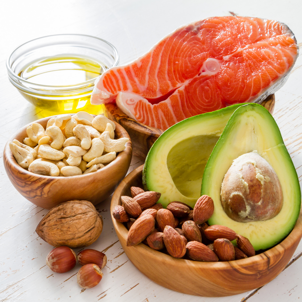 Healthy fats on a table. They include olive oil, salmon, cashews, almonds, avocado and more
