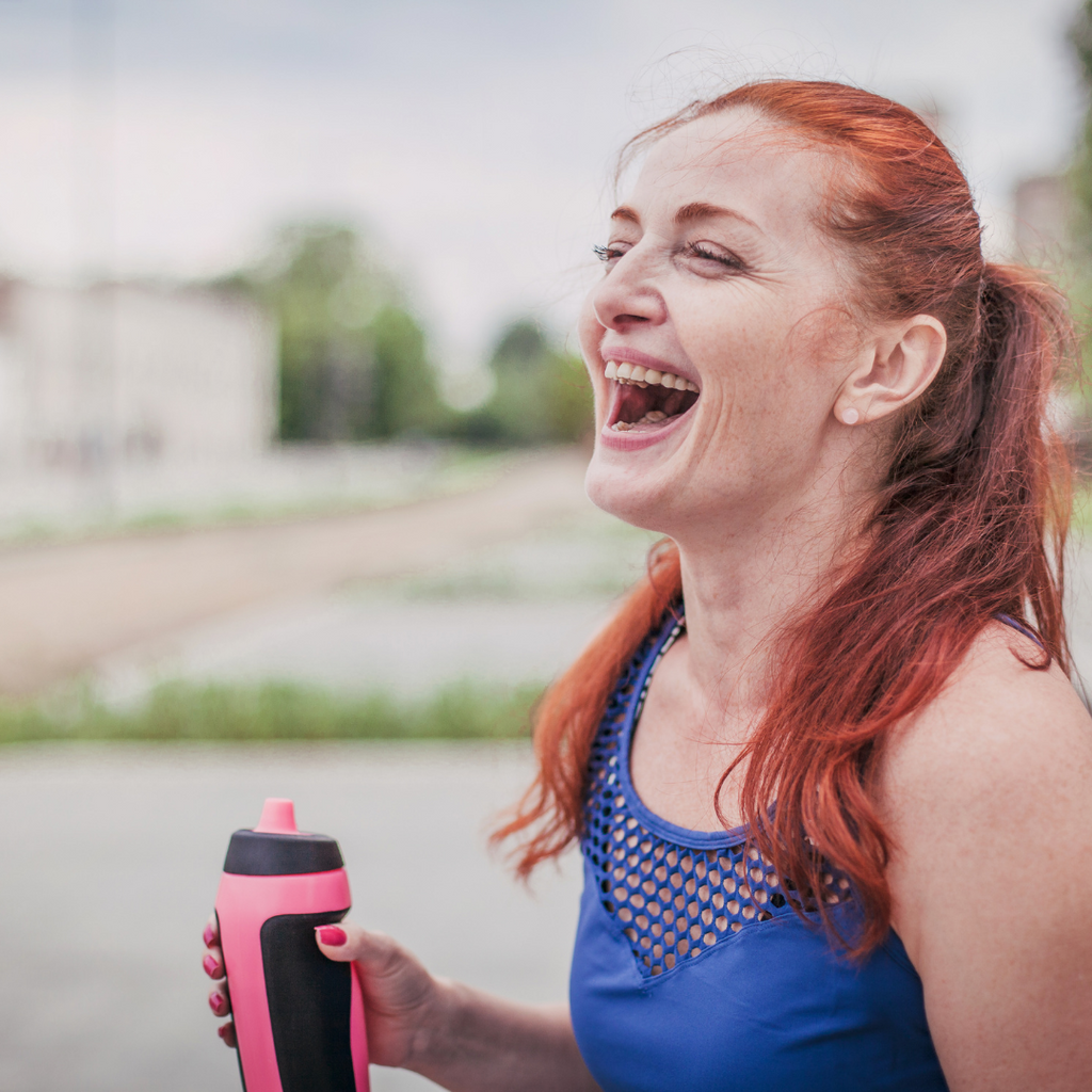 A red haired lady wearing activewear and holding a water bottle. She is laughing.