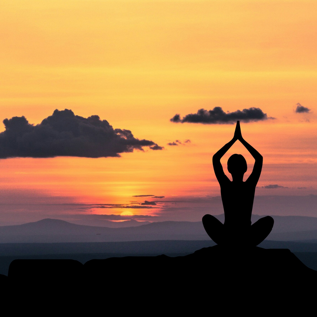 A woman's silhouette. She is doing yoga on a rock, staring into the horizon as the sun is setting.