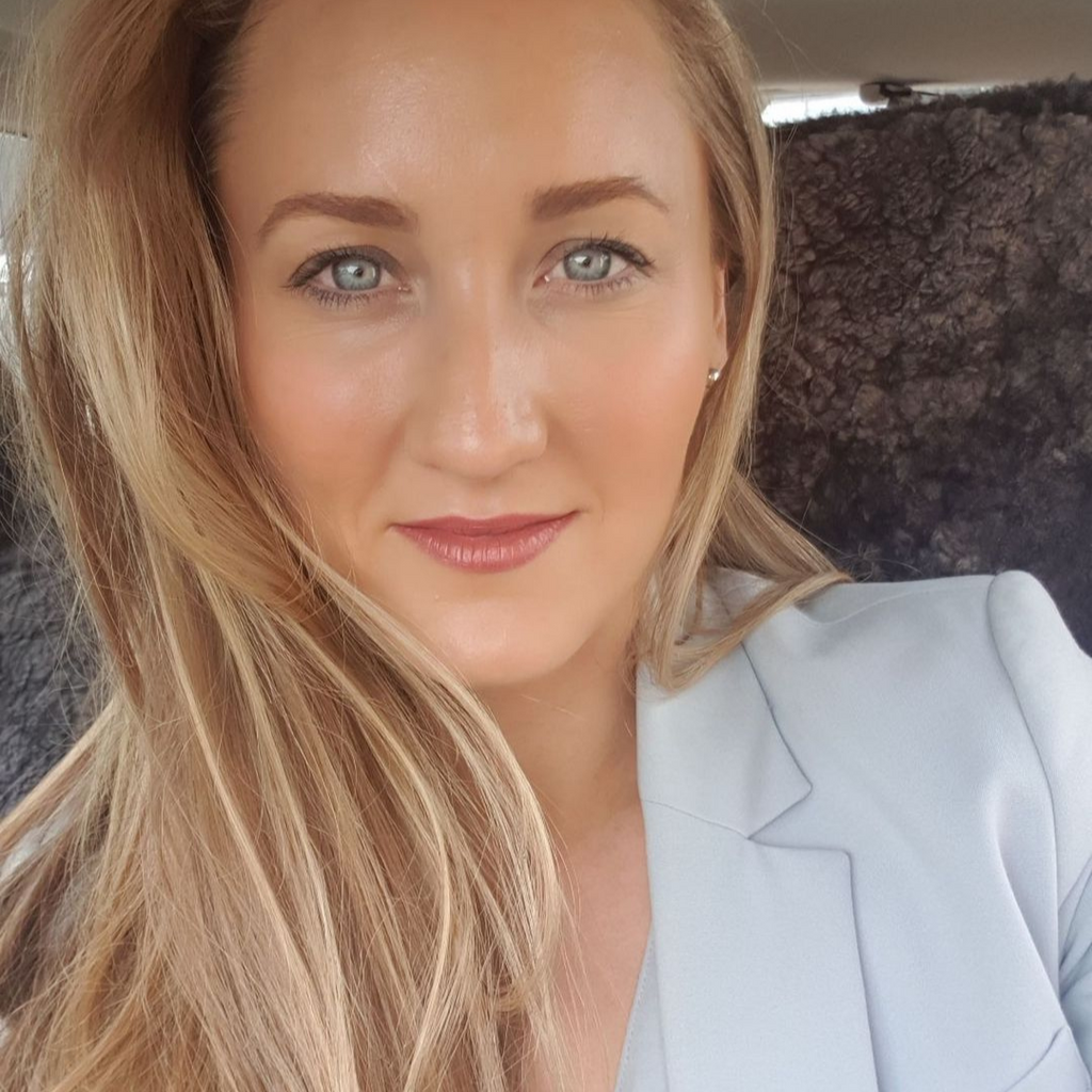 A close up selfie of Lauren. She is sitting down and wearing a baby blue blazer. Her blonde hair is swept to her right side and she is smiling softly at the camera. 