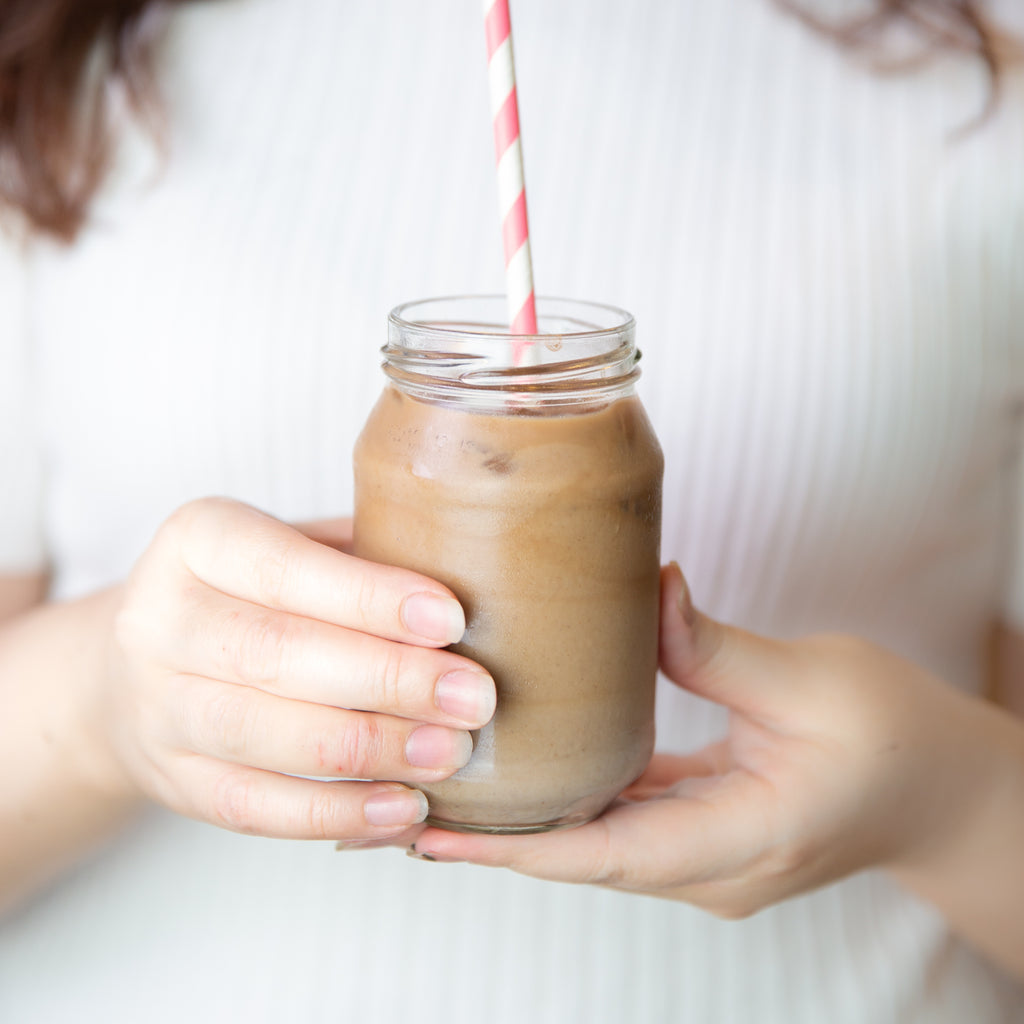 A cacao latte held by female hands. Her head is cut off but she is holding the mason jar delicately. The milk/latte in the jar has a swirl effect as though it had just been mixed together. 