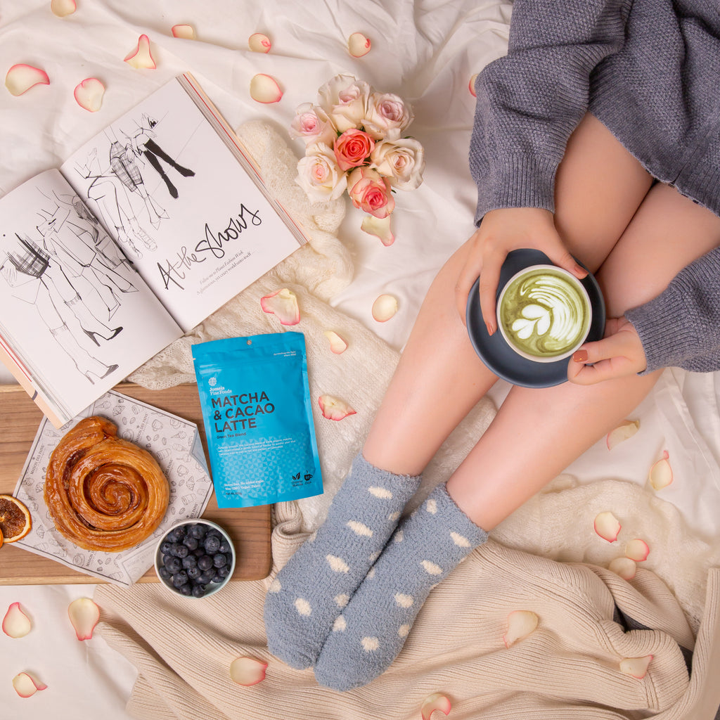 A girl holding a matcha and cacao latte in a seated position. She is wearing an ovesized jumper and grey fuzzy socks. Beside her is a cinnamon scroll and a matcha and cacao latte packet. 