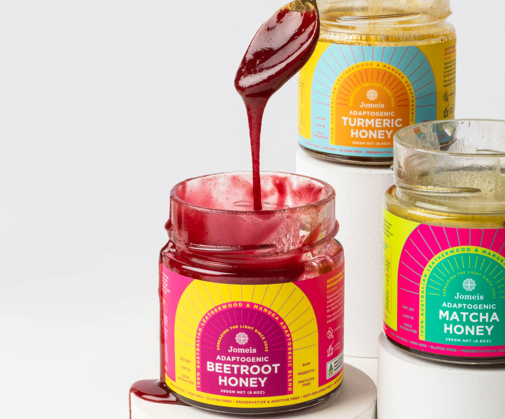 An open beetroot honey with a spoon coming out of it. The spoon is doused in pink beetroot honey which is dripping back into the container. In the background there is an open matcha honey and an open turmeric honey.