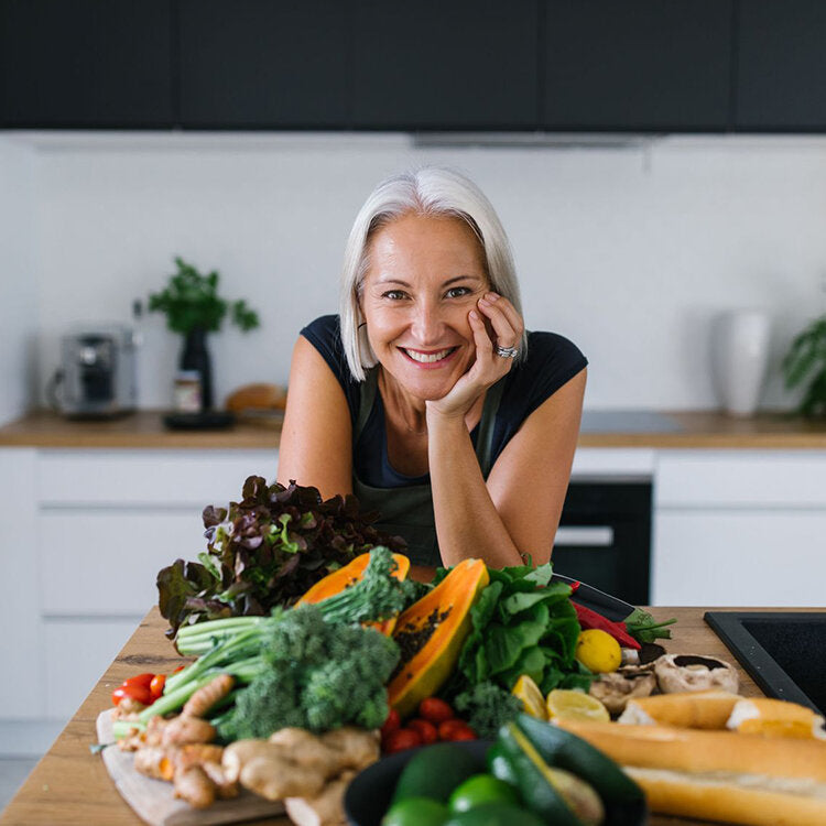 An image of Dani Stevens learning on her kitchen bench. Her head is perched on her hand. In front of her is an array of fruits and veggies including broccoli, spinach, papaya and lime. She is smiling brightly at the camera.