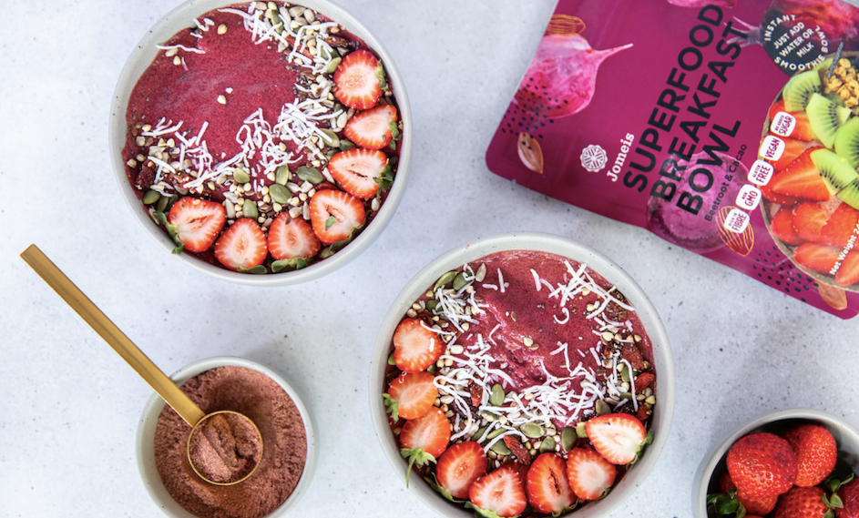 two beetroot and cacao superfood breakfast bowls on a table. Beside them is a beetroot and cacao breakfast bowl packet. The two made bowls are topped with nuts, shaved coconut and strawberries. 