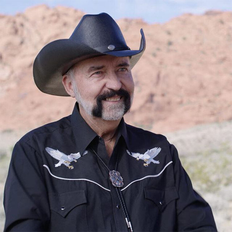 A mid shot image of Don Tolman. He is wearing western attire with a country style short and hat. These are both black. Don is smiling past the camera and it appears he is speaking with someone. Behind him is a dessert-type background. 