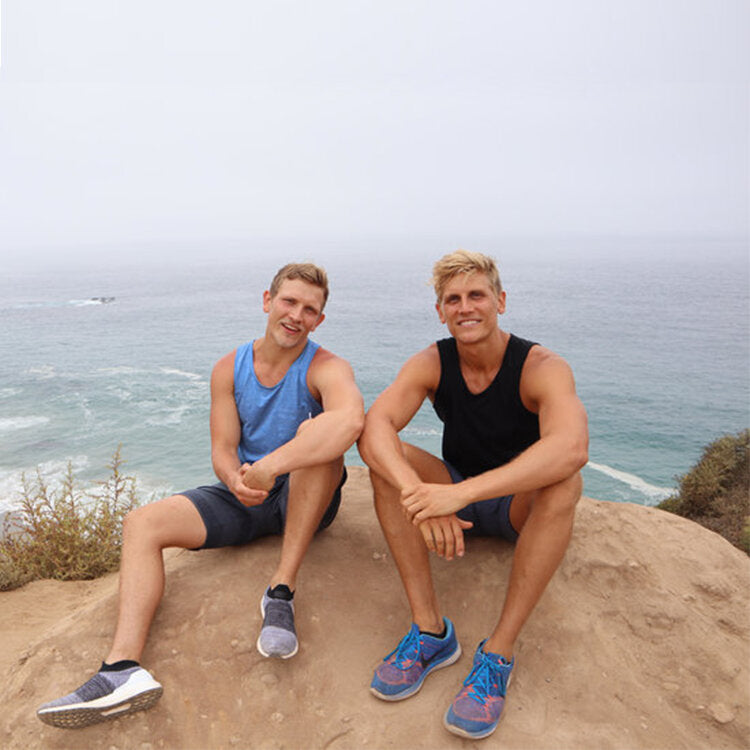 An image of Saxon & Sinclair Fischer-Gray sitting on the edge of a cliff. They are both wearing sports tanks and running shorts. They are facing the camera with the ocean in the background. It is a foggy day. They are both smiling at the camera.