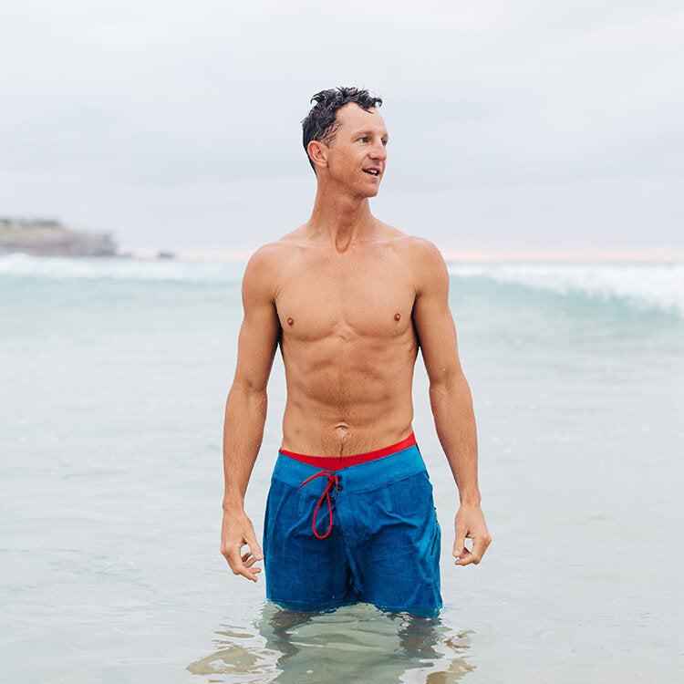 A long shot image of Dean Gladstone standing in the ocean. He is wearing blue board shorts. Dean is staring away from the barrel of the camera and appears to be looking into the ocean. A wave is forming in the distance. 
