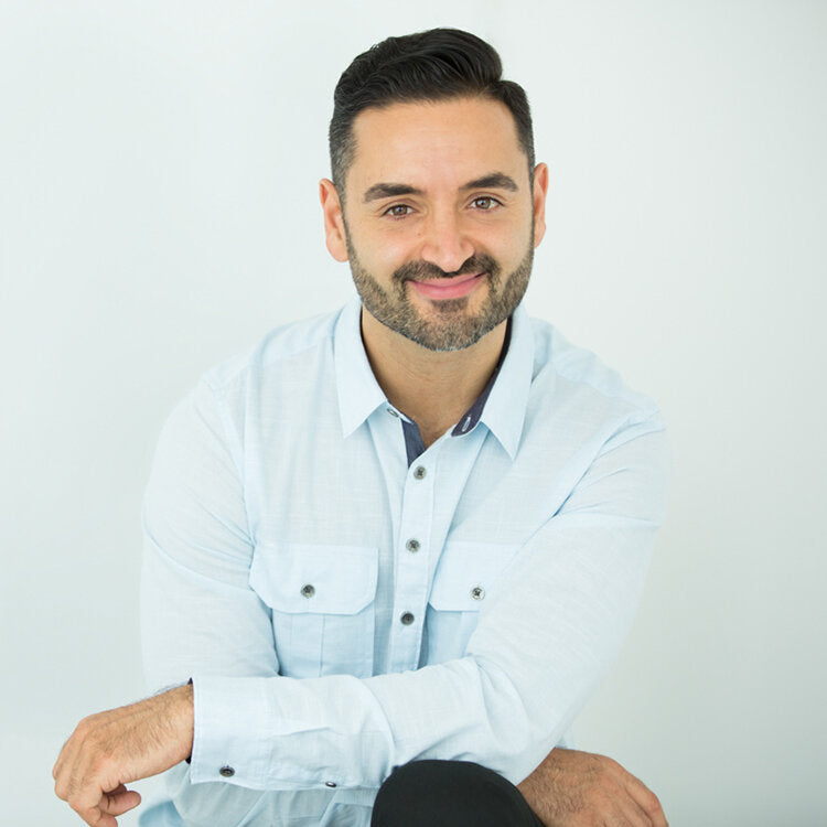 An image of Dr Nima Rahmany. It appears he is sitting on a stool as his legs are crossed over. He is smiling directly at the camera and his arms are folded over his knees. He is wearing a blue button up which works seamlessly with the white background.