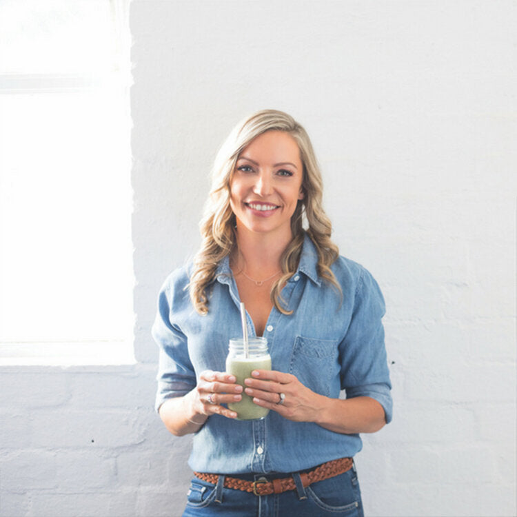 A long shot image of Steph Lowe. She is wearing a blue button up shirt and holding a matcha latte with both of her hands. She is smiling at the camera. Her hair is worn loosely over her shoulders. There is a while brick background behind her. 