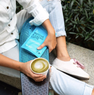 An image of a lady's legs as she is seated on a ledge. She is holding a black and blue skateboard across her legs. In her left hand, she also holds ablue matcha and cacao latte packet. In her right, she is holding a matcha and cacao latte.