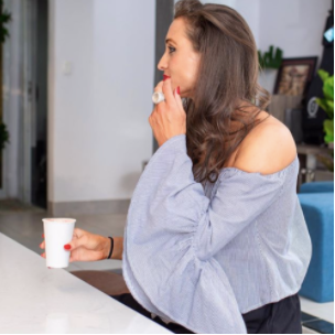 Jomeis Fine Foods founder, Vicki Nguyen sitting at a kitchen bench. She is holding a nutritional latte in her right hand and has her left hand resting on her lips. She is wearing a flowy blue shirt and black pants. 