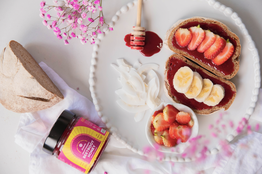 An adaptogenic beetroot honey is at the bottom right of the screen. Above it is half a sourdough loaf and to the right is a plate with two pieces of sourdough. The beetroot honey is spread on the sourdough and topped with bananas and strawberries. 