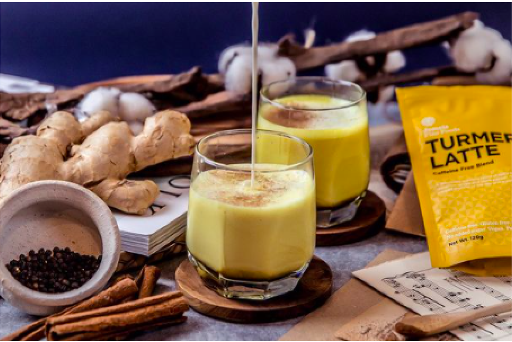 A turmeric latte being poured. Behind it is another turmeric latte and to the right is a turmeric latte packet. the liquid being poured is yellow and it has been topped with cinnamon, this can be seen as the glasses in use are clear.