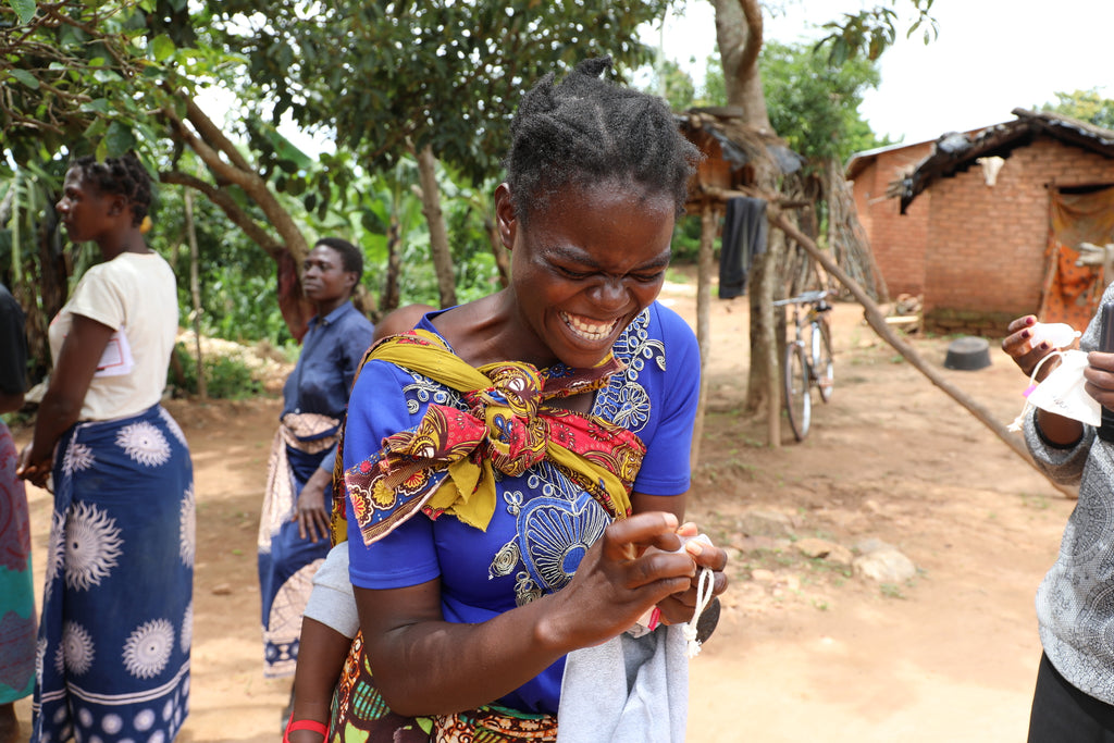 A young African girl wearing a blue dress. She is smiling excitedly at her hands. In her hands she is holding a menstrual cup which has just been donated to her through The Cova Project. She is outdoors and there are people in the background. 