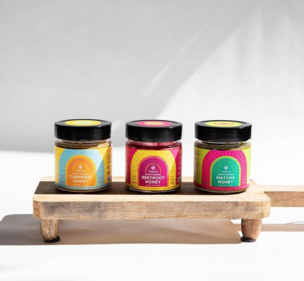 Three containers of adaptogenic honey sitting on a chopping board. From left to right they are, yellow turmeric honey, pink beetroot honey, green matcha honey. All honey containers are sealed. 