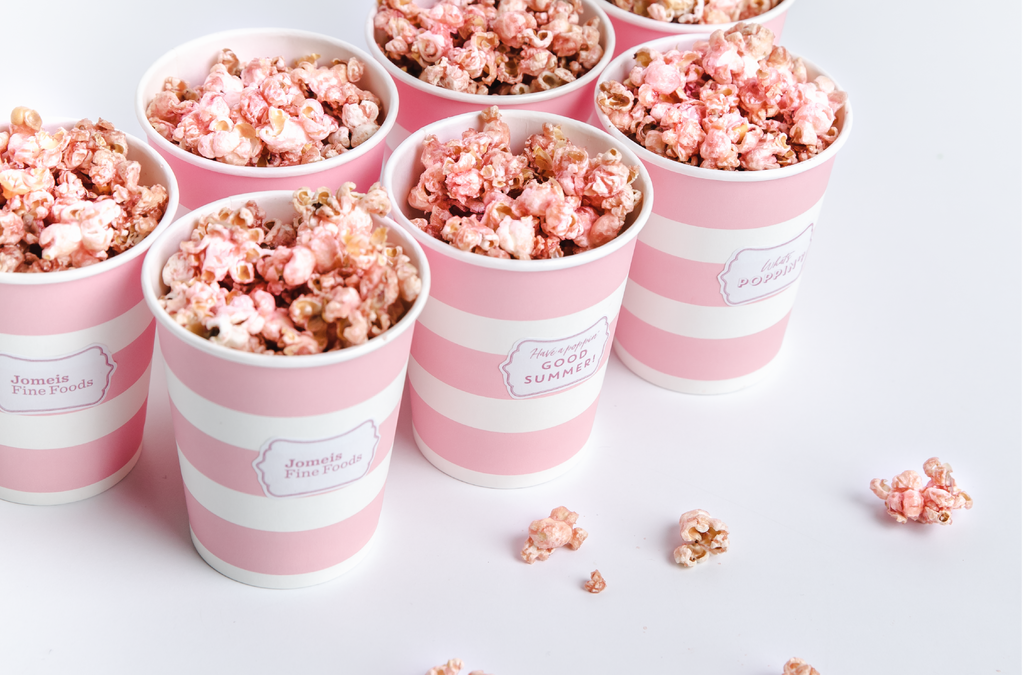 seven pink and white striped cups that are filled with popcorn. The popcorn has a light, beetroot dust on it making them a baby pink colour. All the cups are tucked in the top left corner of the image. In the bottom right, there is some spilt popcorn