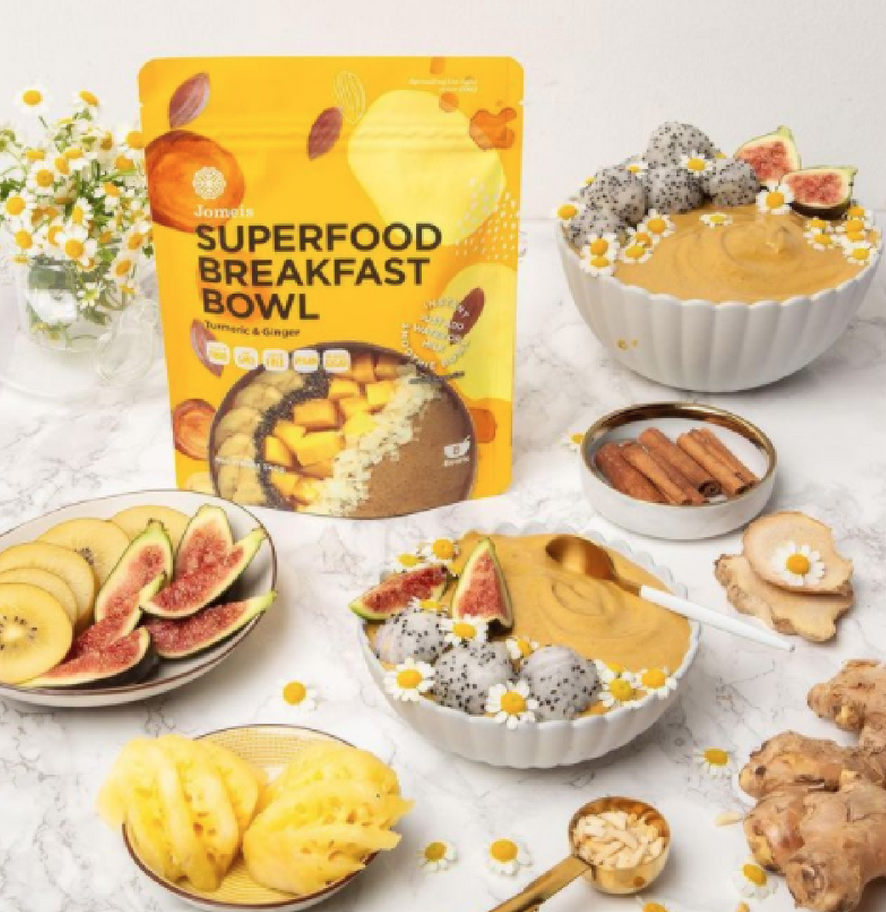 Yellow superfood breakfast bowl packet surrounded by yellow fruits and a made up breakfast bowls. The breakfast bowls are paired with fig and dragon fruit. The background is marble and there are flowers in the distance. 