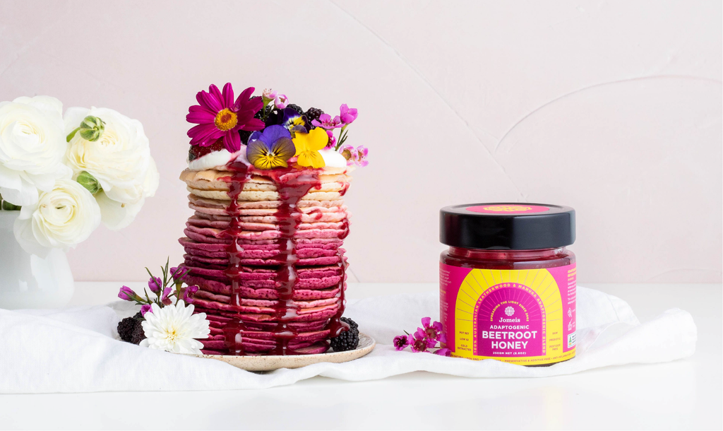 A stack of pink beetroot pancakes topped with edible flowers. To their right is a pink beetroot honey tub. The pancakes have the honey dripping off of them. The plate of pancakes and the honey are resting on a white sheet to create an elegant feel