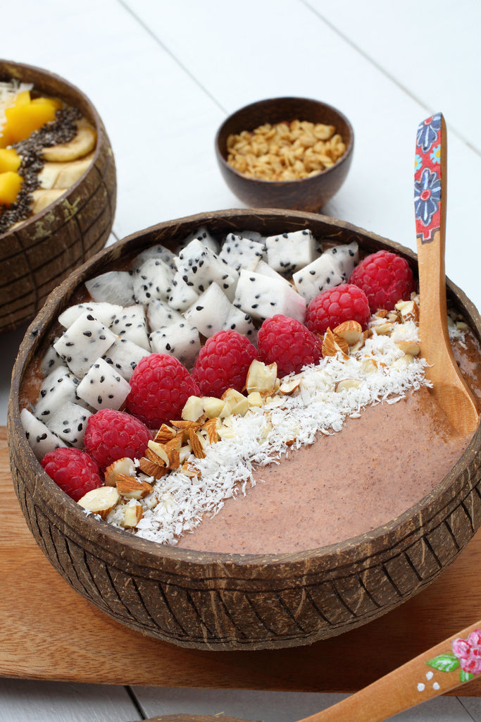 A cacao and coconut superfood breakfast bowl topped with coconut, fruit and nuts. It is in a coconut bowl and a wooden spoon is  sticking up from it. 