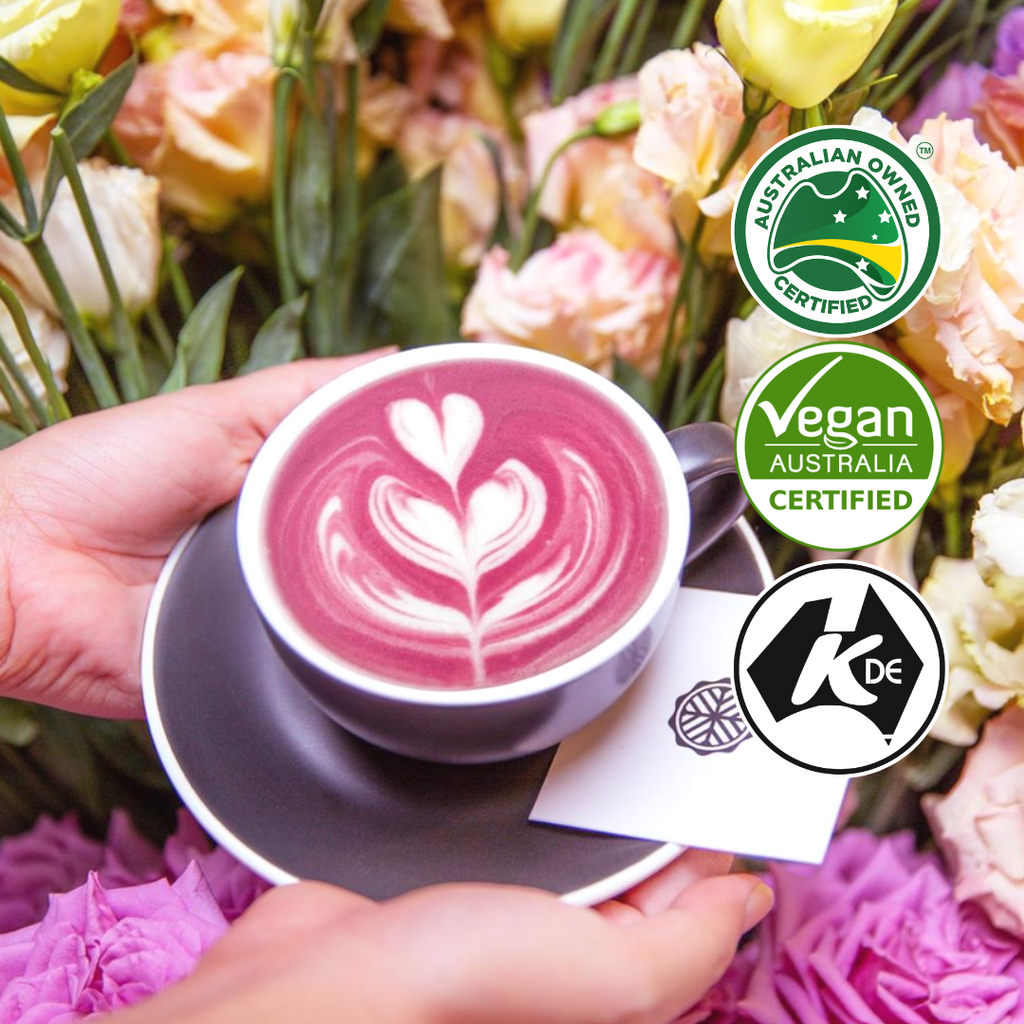 A cup of beetroot latte with detailed latte art. It is held delicately with two hands. There is a bouquet of flowers accompanying the background.
